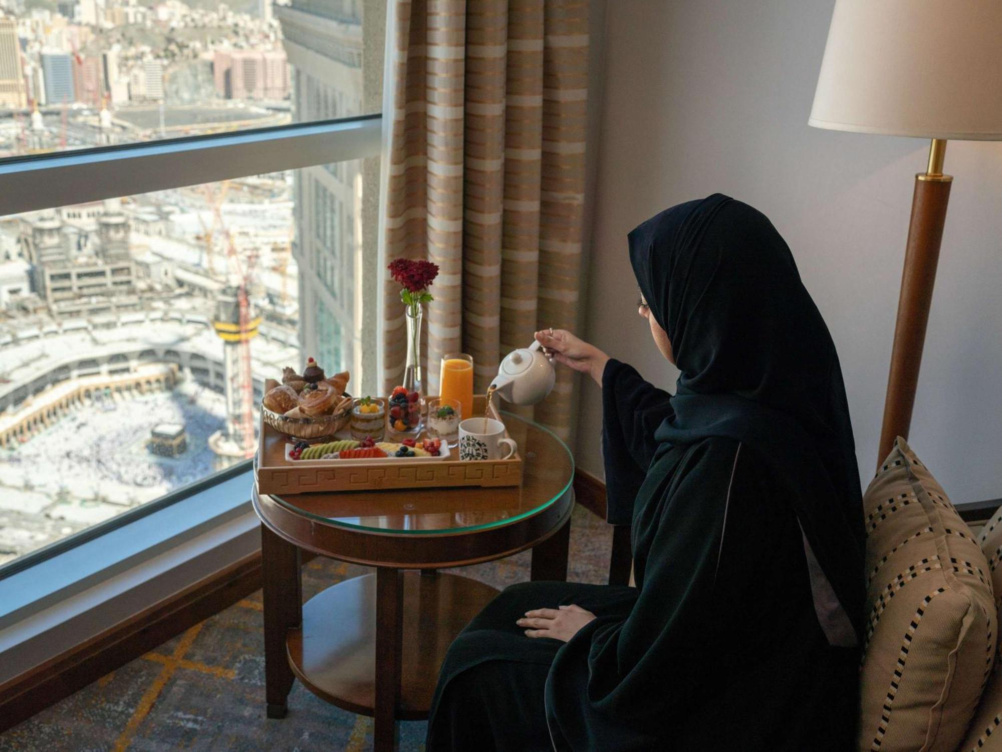 Swissotel Al Maqam Makkah Mekka Buitenkant foto The image depicts a woman wearing a black abaya, sitting at a table near a large window. She is pouring tea from a teapot into a cup. The table has a beautifully arranged tray with various food items, including pastries and fruits, as well as a glass