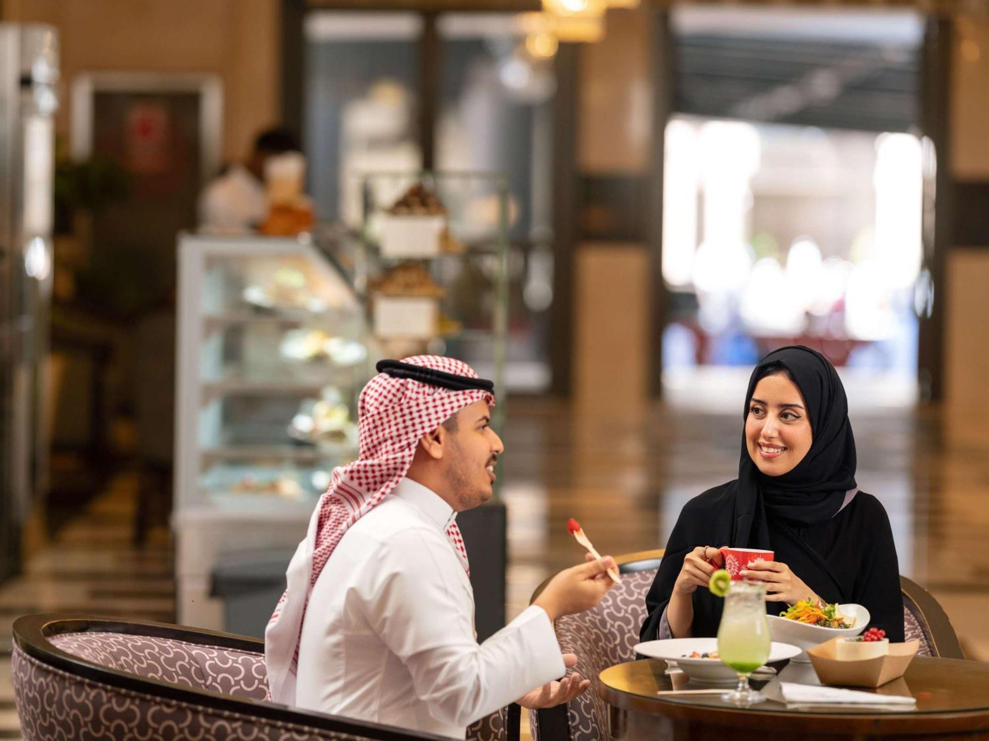 Swissotel Al Maqam Makkah Mekka Buitenkant foto The photo features a man and a woman engaged in a conversation at a dining table. The man is wearing traditional attire with a white thobe and a headscarf, while the woman is dressed in a black abaya and headscarf. They appear to be enjoying a meal t