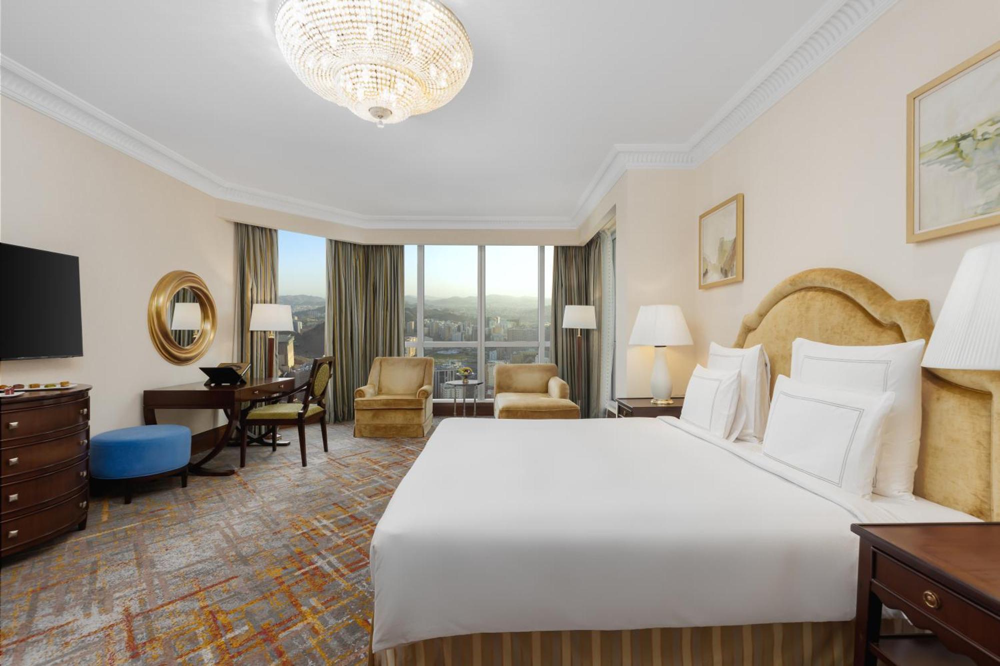 Swissotel Al Maqam Makkah Mekka Buitenkant foto The photo shows a well-furnished hotel room with a modern and elegant design. It features a large bed with white linens and decorative pillows. There are two armchairs nearby, a small round table, and a writing desk with a chair. A television is moun