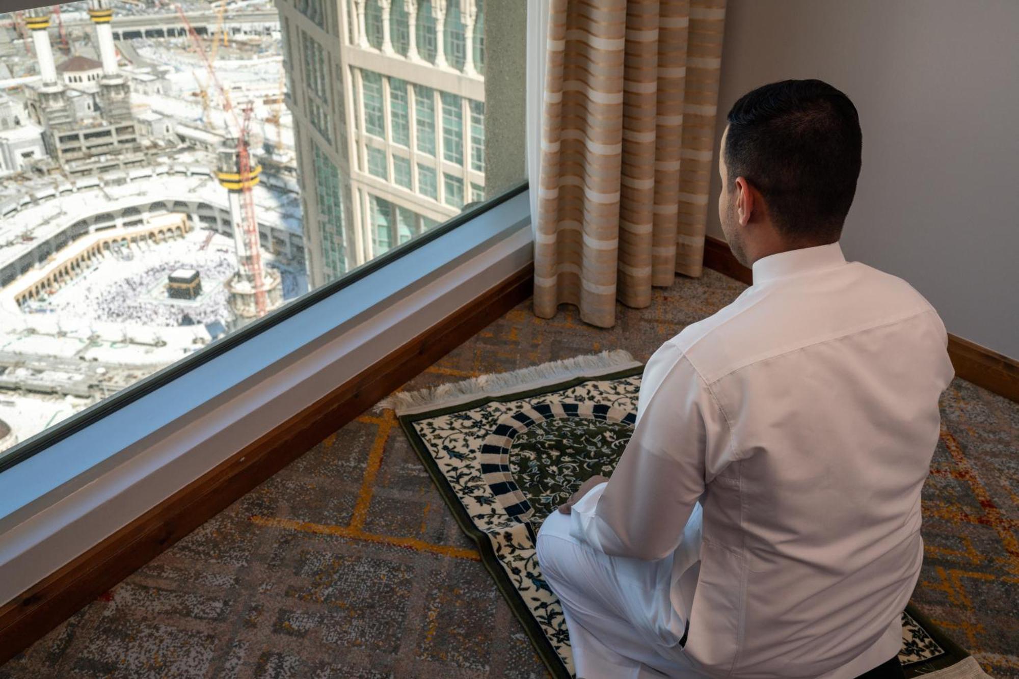 Swissotel Al Maqam Makkah Mekka Buitenkant foto The photo depicts a person sitting on a prayer mat, facing a large window. Through the window, a view of a bustling area is visible, likely near a significant religious site, as suggested by the presence of a mosque or similar structure outside. The 