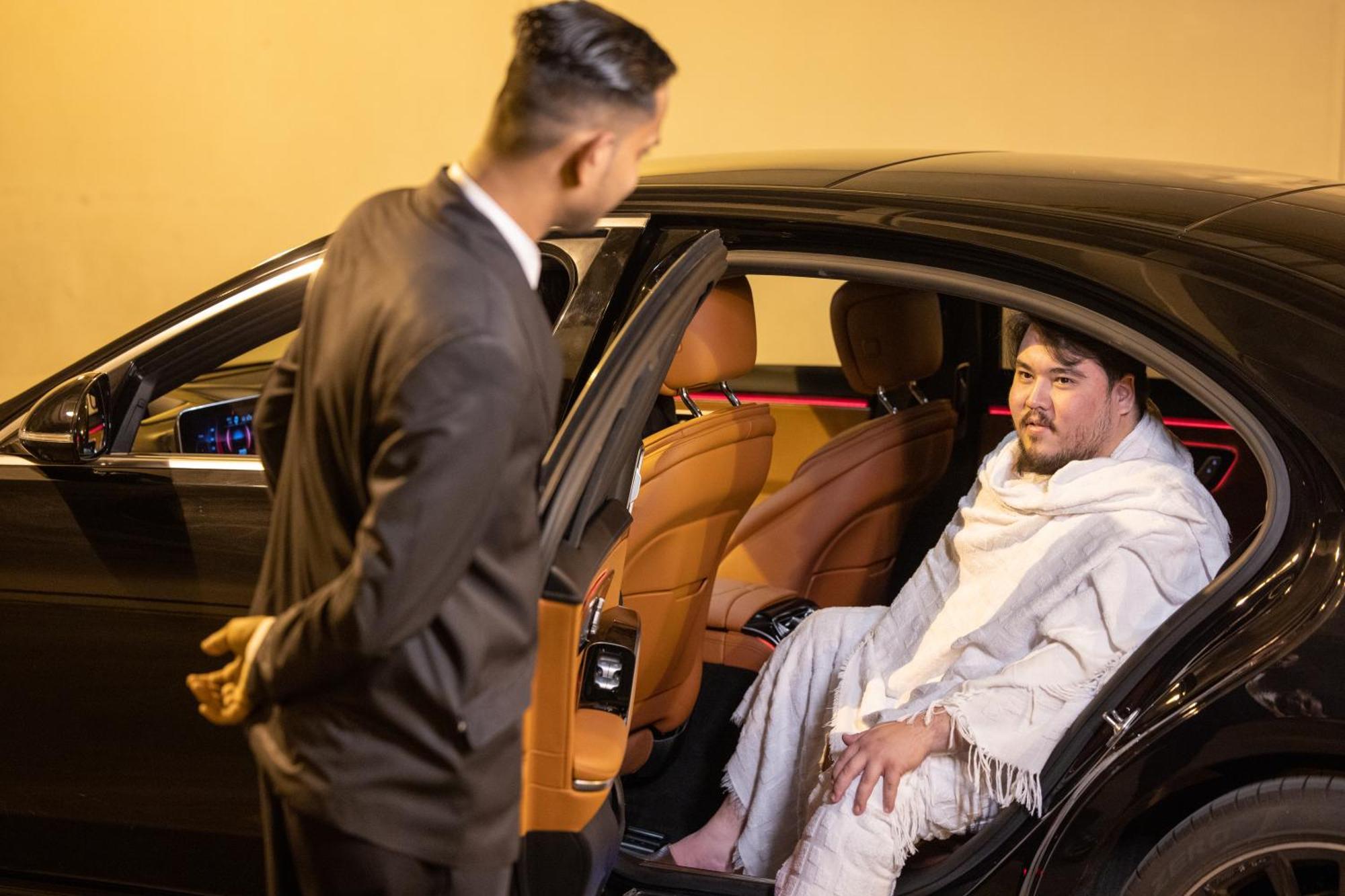Swissotel Al Maqam Makkah Mekka Buitenkant foto The image shows a scene outside a car at night. One person, dressed in a formal black suit, is standing next to an open car door, seemingly talking to the other person inside the car. The person inside the car appears to be wearing a white blanket or