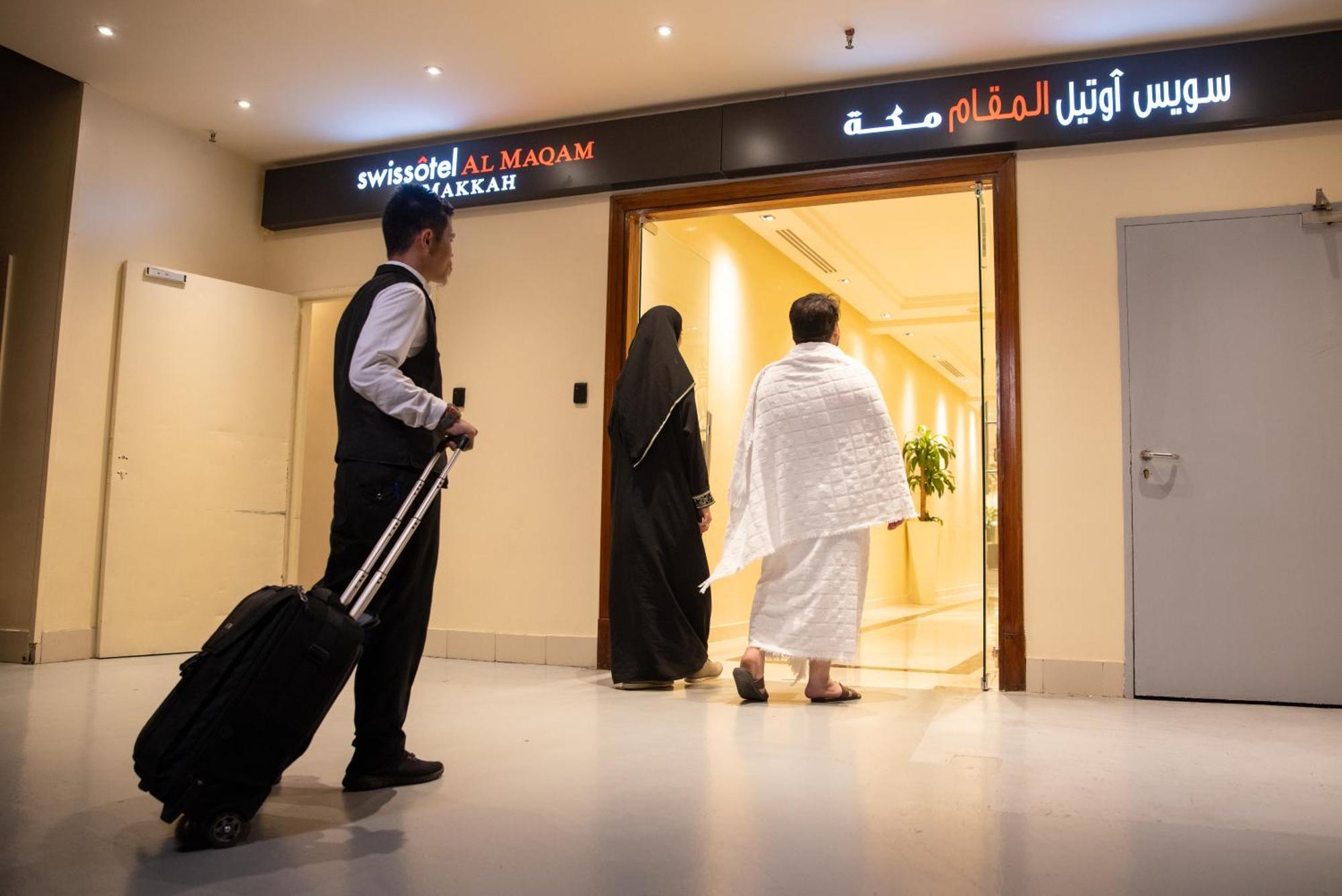 Swissotel Al Maqam Makkah Mekka Buitenkant foto The photo shows a hotel entrance, specifically for the "Swissotel Al Maqam Makkah," as indicated by the sign. In the scene, a man in a formal outfit carrying a suitcase waits nearby, while two individuals, one in traditional clothing (possibly a woma