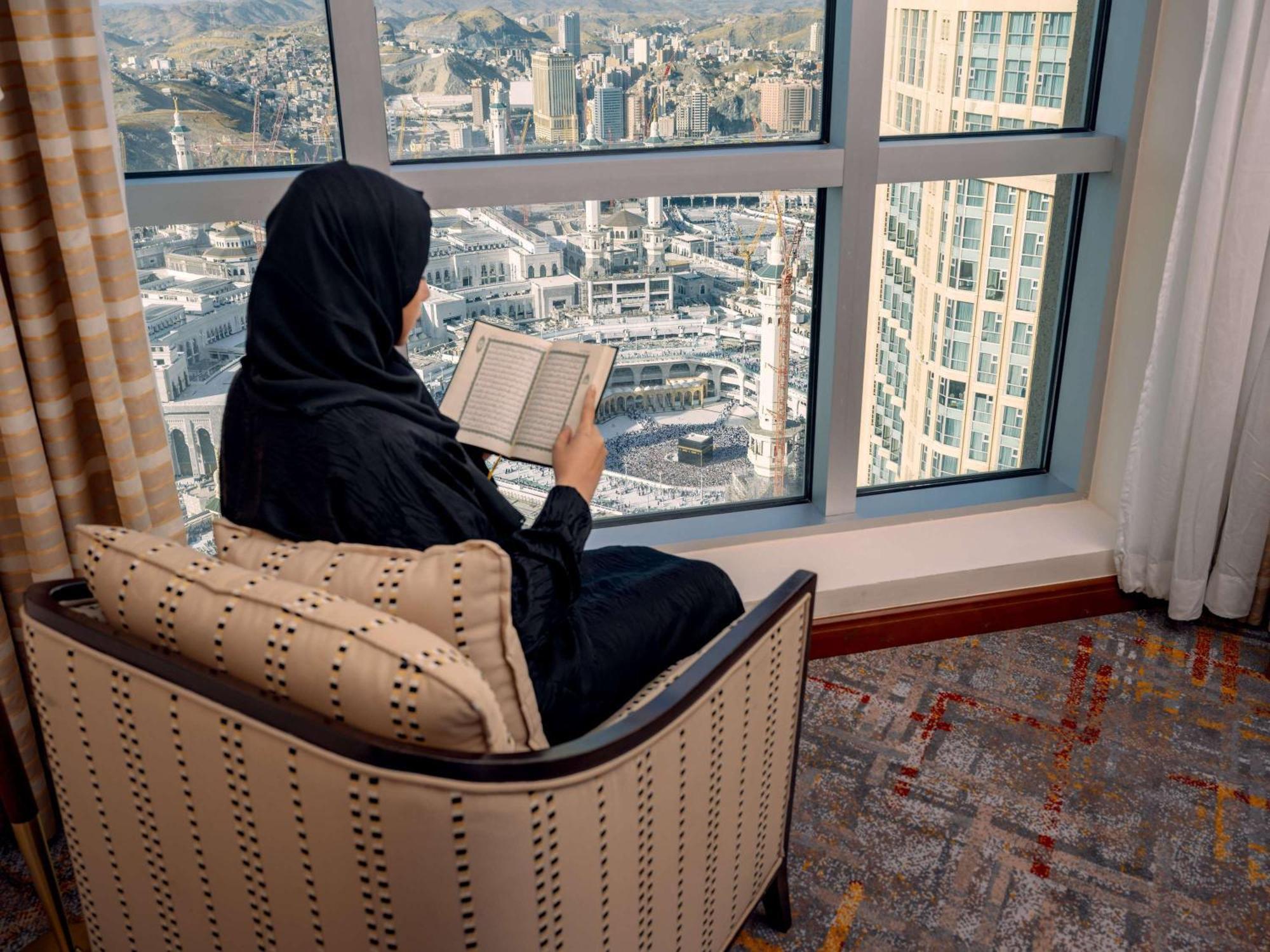 Swissotel Al Maqam Makkah Mekka Buitenkant foto The photo depicts a woman seated in a comfortable chair by a large window. She is wearing a traditional black garment and is focused on reading a book. Outside the window, there is a view of a cityscape, possibly a metropolitan area, with various bui