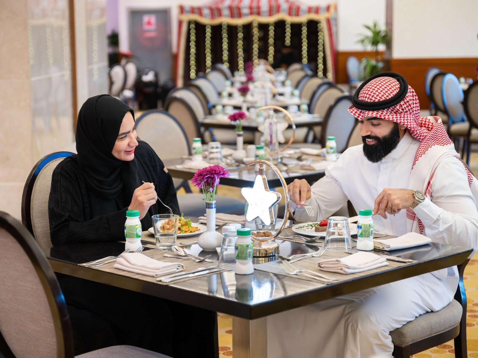 Swissotel Al Maqam Makkah Mekka Buitenkant foto The photo shows a couple dining at a restaurant. The man is wearing traditional attire, including a white thobe and a red and white checkered headscarf, while the woman is dressed in a black abaya and a headscarf. They are engaged in a pleasant conve