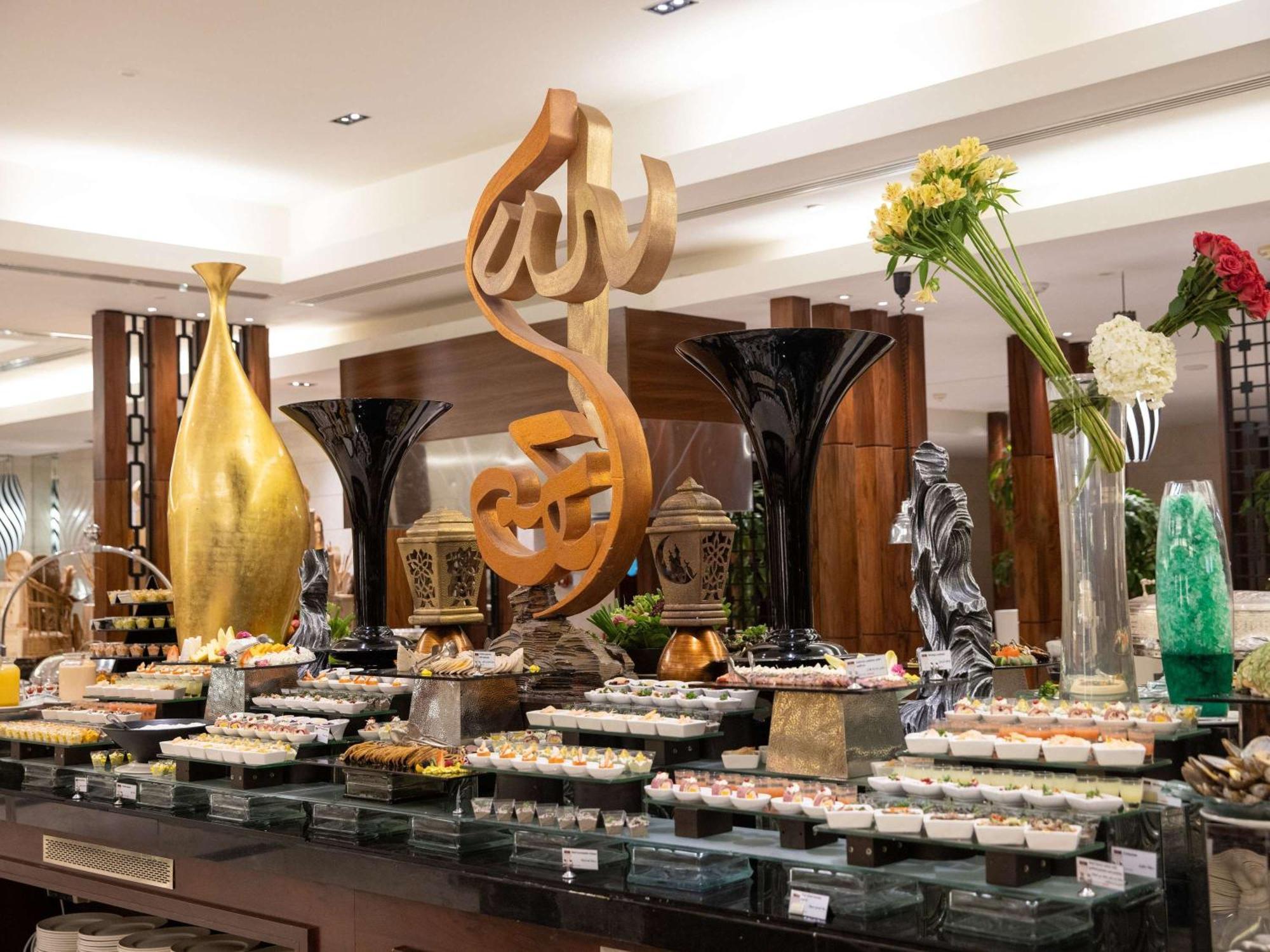 Swissotel Al Maqam Makkah Mekka Buitenkant foto The photo shows a beautifully arranged buffet display in a restaurant. In the center, there is a large decorative piece featuring Arabic calligraphy. Surrounding it are various elegant food presentations, including trays of desserts, likely reflectin