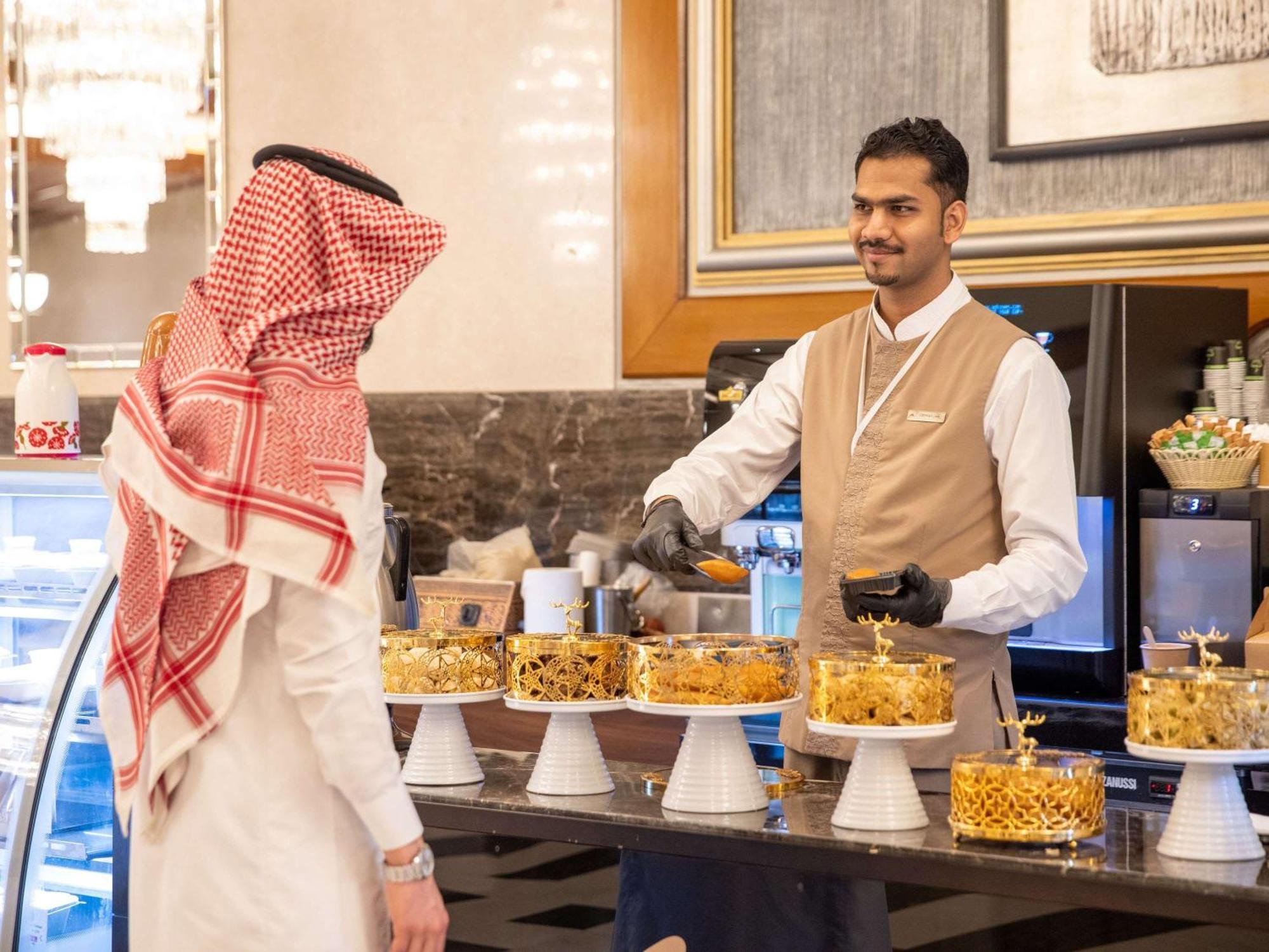 Swissotel Al Maqam Makkah Mekka Buitenkant foto The photo depicts a scene in a bakery or dessert shop. There is a display of various beautifully decorated cakes arranged on stands. A man wearing traditional attire, possibly a thobe and a headscarf, is interacting with a staff member who is smiling