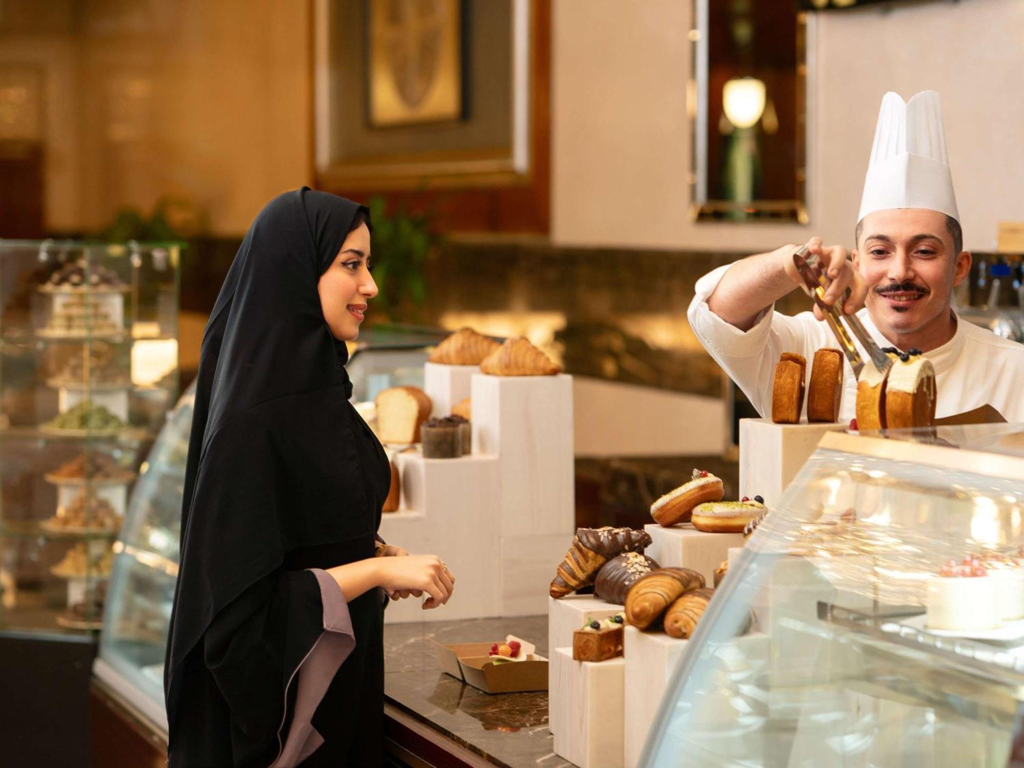 Swissotel Al Maqam Makkah Mekka Buitenkant foto The photo shows a bakery or pastry shop scene. On the left, a woman is wearing a black abaya and appears to be interacting with a male baker. The baker, dressed in a white chef's hat and uniform, is behind a display counter filled with various baked 