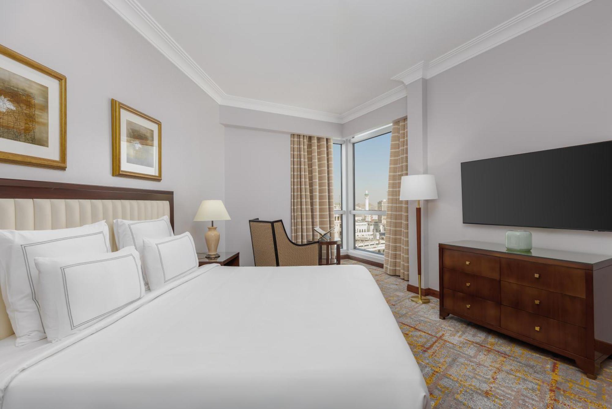 Swissotel Al Maqam Makkah Mekka Buitenkant foto The photo shows a hotel room featuring a neatly made bed with white linens and several decorative pillows. There's a comfortable chair positioned near a window, which has light curtains allowing natural light to enter. A bedside lamp and framed artwo